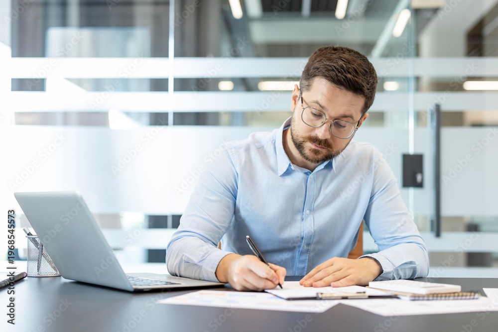Naklejka premium Businessman wearing glasses and a light blue shirt concentrating on writing notes on a document while working at a desk, with a laptop and various papers in a modern office environment