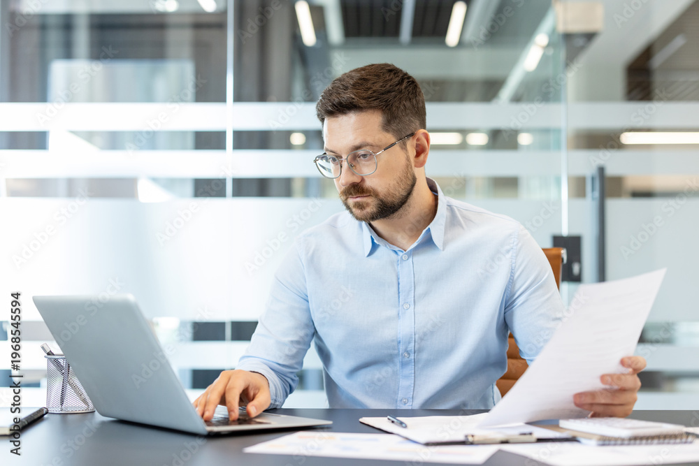 Fototapeta premium Businessman wearing eyeglasses and a blue shirt, concentrating on his laptop and carefully reviewing paperwork while sitting at a desk in a modern corporate office environment
