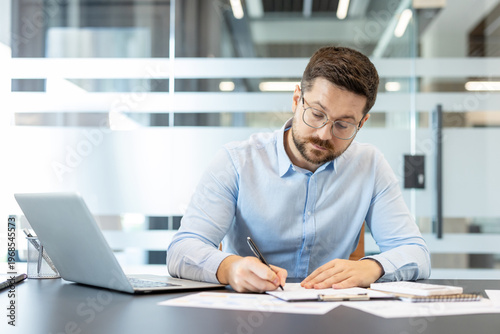 Businessman wearing glasses and a light blue shirt concentrating on writing notes on a document while working at a desk, with a laptop and various papers in a modern office environment