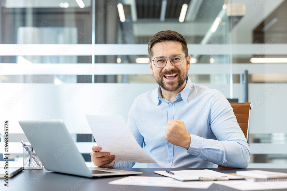 Obraz premium Happy businessman cheering with a fist pump and a wide smile, holding official document in hand while sitting at a desk with a laptop and paperwork, celebrating good news and career achievement
