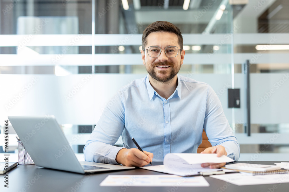 Fototapeta premium Young man in glasses and blue shirt doing paperwork at his desk, holding a pen and looking at camera with a cheerful expression in a modern corporate workspace setting