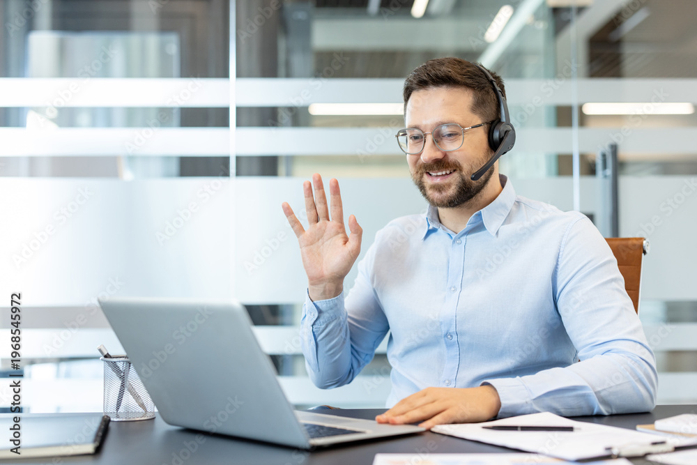 Obraz premium Man wearing a headset and spectacles, smiling and waving hand to laptop screen, participating in a remote work virtual meeting from his modern office desk