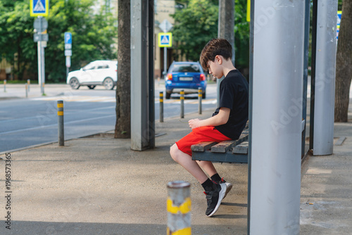 Young boy waiting alone at urban bus stop sitting on a bench at a bus stop feeling sad and lonely