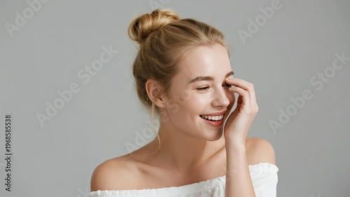 A smiling woman with blonde hair in a bun, wearing an off-shoulder white top, touching her face against a neutral background.