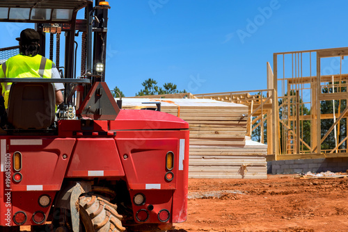 Forklift operator moves stacks of lumber on construction site where workers build new structures outside.