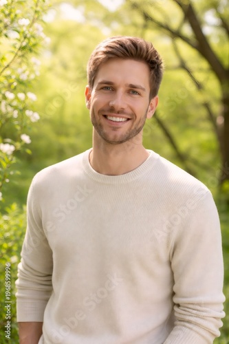 Young man smiling confidently, standing in a vibrant green natural setting with blooming trees, expressing happiness, positivity, and a carefree lifestyle