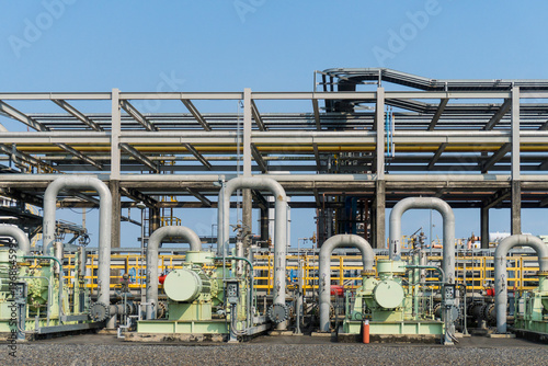 Industrial plant with pipes and machinery under clear blue sky. Workers in safety gear walk among equipment. Scene shows active manufacturing or energy production. Suitable for engineering