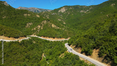 Aerial view of a campervan traveling, driving the iconic SH75 mountain road in Albania, winding through the rugged landscape above the vibrant turquoise waters of the Vjosa River in the summer sun.