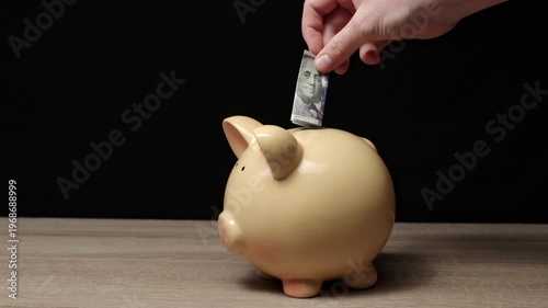 Hand Inserting Bill Into Piggy Bank On Wooden Table Closeup Against Black Background Showing Deliberate