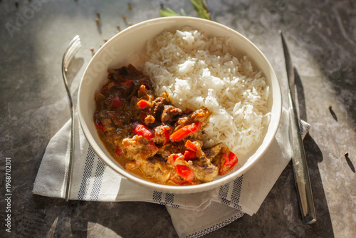 Hearty beef stew with red bell peppers and fluffy white rice served in a bowl on a dark marble counter in bright sunlight