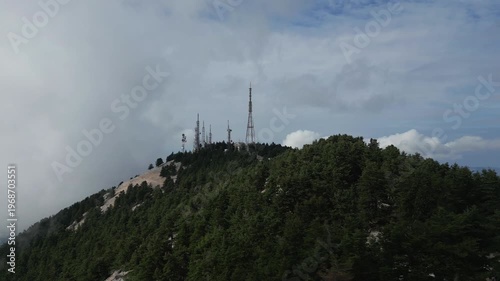 Towers rise above a forested mountainside, poking through clouds under a hazy sky.
