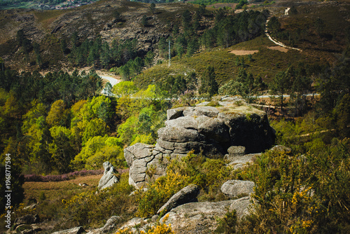 Rocky landscape in Peneda Geres National Park with granite boulders, green hills, forest patches, and a winding road in the background.