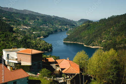 View of Cavado river valley in Peneda Geres with forested hills, calm water, and houses with red tile roofs in the foreground.