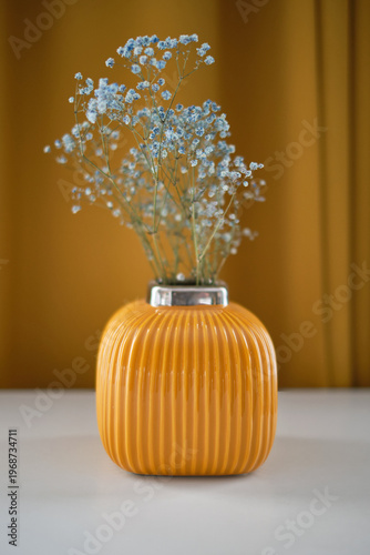 A yellow vase with dried flowers on a white table against a background of yellow curtains. Selective focus
