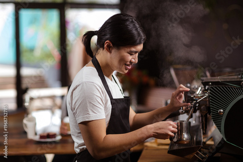 Asian barista in twenties operating espresso machine at coffee shop, frothing milk with pitcher