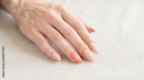 A close-up of a woman's hand with long, almond-shaped nails. The nails are painted in nude and coral colors. The hand rests on a light fabric surface.