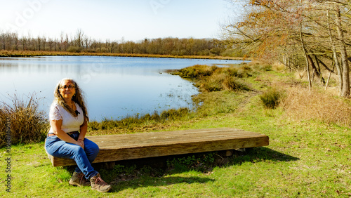 Smiling, serene adult woman sitting on wooden bench with a lake in background in the Tosch-Langeren nature reserve, surrounded by bare trees, winter day in Maaseik, Belgium. Space for text