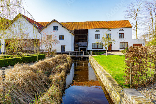 Canal between green grass, dry weeds and path with a watermill in background, farm with white walls and gabled roof, reflection on surface of the water, sunny winter day in Maaseik, Belgium