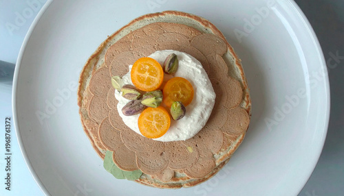 Stack of fluffy pancakes with whipped cream, kumquats, and pistachios in sunlight