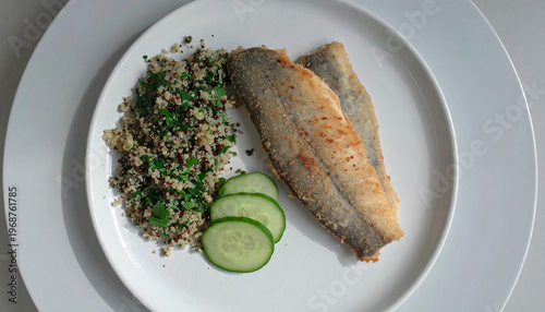 Fried fish fillets served with quinoa salad and cucumber slices on a white dish