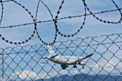 Jet Landing: White Plane, Barbed Wire and Cloudy Sky. White Passenger Plane Landing at Airport over Barbed Wire Fence under Cloudy Sky