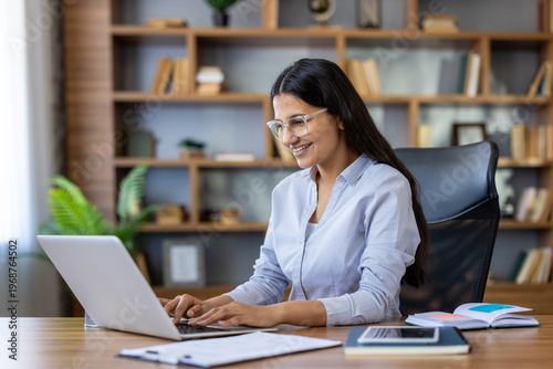 Young indian woman wearing glasses cheerfully working on a laptop at her desk in a contemporary home office or library setting, focusing on her online tasks