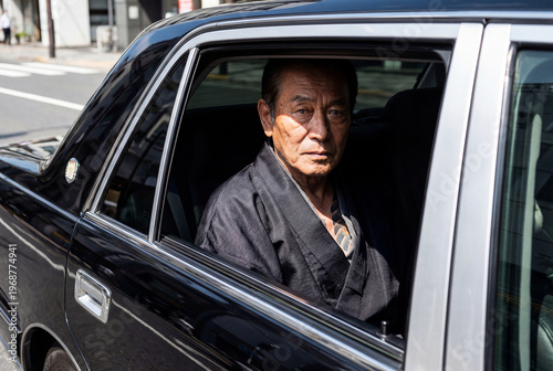 Stern senior Japanese man wearing traditional dark clothing sitting in the back seat of a black car, looking out the open window on a city street.