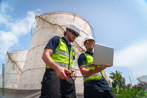 Industrial workers in safety vests and hard hats review data on a laptop near storage tanks