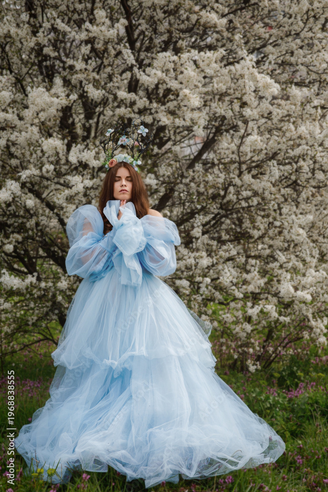 Fototapeta premium young woman in blue dress standing under blooming tree with floral headpiece, soft spring light, delicate white blossoms, dreamy romantic garden portrait