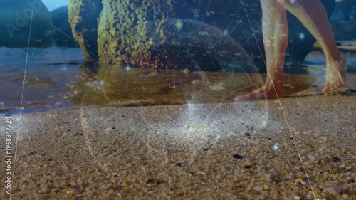 Animation of globe with rays against caucasian man walking barefoot at sunny beach