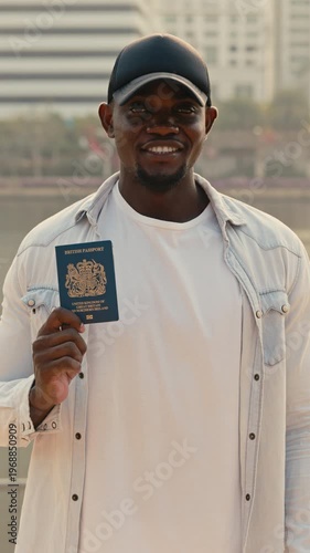 Portrait of African American man in cap showing British passport looking at camera. Smiling guy in denim shirt holds document confirming citizenship. Male stands on city embankment
