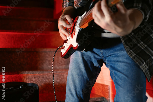 Close-up of Electric Guitar Player with Dramatic Red Lighting. Musician hands playing a black and white electric guitar. Atmosphere with vibrant red backlight, featuring an amplifier in the background