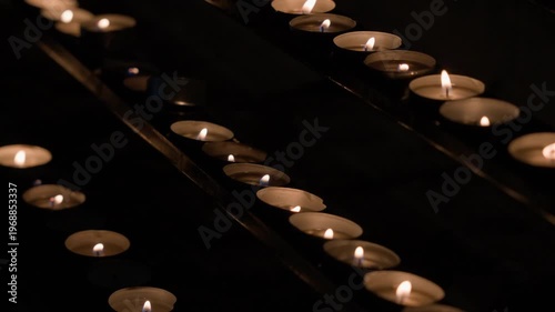 Close-up view of many candles arranged diagonally with shallow depth of field. Static camera captures warm reflections and soft glowing flames in dark environment.