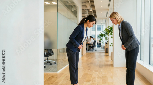 Two businesswomen bowing to each other in a modern office hallway. Diverse female colleagues in dark suits exchanging a respectful greeting. Corporate business etiquette and international partnership