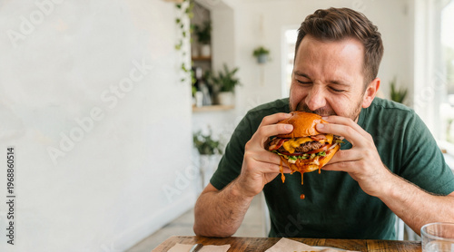 Hungry man taking a big bite of a messy cheeseburger. Male enjoying a huge delicious fast food burger with dripping cheese and bacon in a cafe