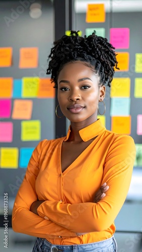 Portrait of a woman with curly hair and arms crossed, wearing an orange blouse. Background has colourful sticky notes on glass