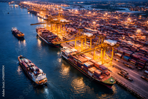 Aerial view of illuminated container port with cargo ships, cranes and stacked containers showing global shipping and logistics operations at night.