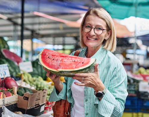 A woman enjoys selecting ripe tomatoes at a bustling outdoor farmers market. Colorful fruits and vegetables surround her as she smiles at the freshness of the produce