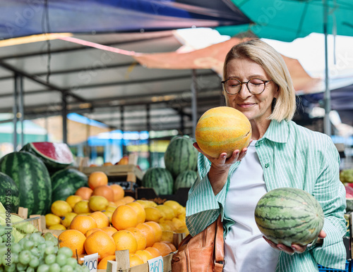 A woman enjoys selecting ripe tomatoes at a bustling outdoor farmers market. Colorful fruits and vegetables surround her as she smiles at the freshness of the produce