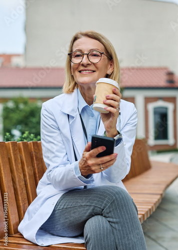 Portrait of a mature older female woman doctor or nurse and  health care worker using a smartphone mobile phone  in front of hospital or clinic outside