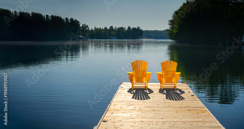 Rear view of two yellow Adirondack chairs on a dock at sunrise in Muskoka Ontario Canada, long shadows on calm glassy lake, cottage country, no people.