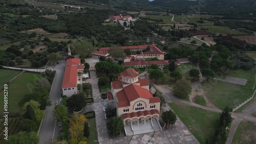Aerial view of a monastery complex nestled among olive groves and rolling hills, featuring a central church with a red dome.