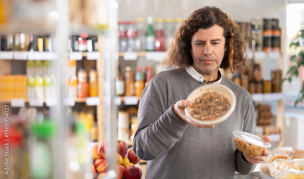 Fototapeta premium Adult man holds a package of ready-made yakisoba noodles, reading the ingredients and looking at the expiration date. European man buys cooked food in a supermarket