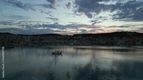A solitary sailboat drifts on calm waters as dusk settles over a coastal town, with a soft, cloudy sky reflecting in the sea.