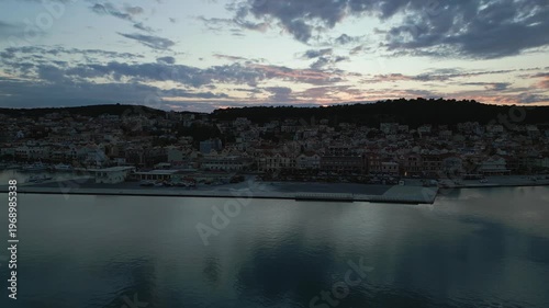 A coastal town at dusk, with boats in the harbor and buildings nestled against a tree-covered hill under a colorful sky.