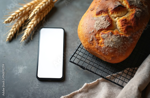 Freshly baked bread sits on cooling rack next to smartphone with blank screen. Wheat stalks nearby. Natural light creates soft shadows on table. Perfect for food bloggers and recipe apps.
