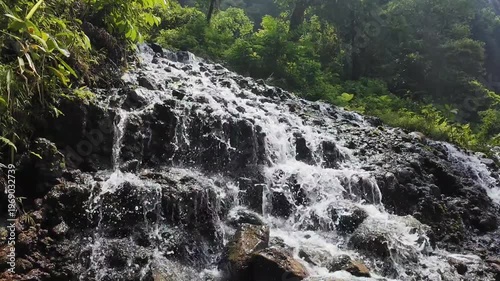 A beautiful natural landscape featuring a majestic waterfall cascade flowing over mountain rocks and stones through a lush green summer forest stream