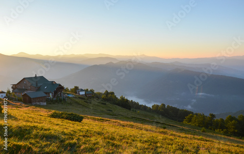 Peaceful mountain cabin sits on grassy slope at sunrise. Layers of misty, blue toned ridges stretch into the distance under pale sky, while golden light illuminates the foreground meadow. Carpathians