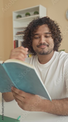 Wallpaper Mural Young indian man reading book at a library table, hand turning page and hand to chin gesture by bookshelf; thoughtful focus. Torontodigital.ca