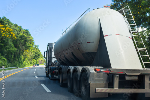 Fuel tanker truck driving on highway surrounded by lush green forest under blue sky, representing logistics and transportation in Brazil.